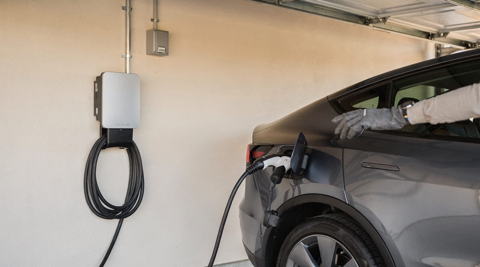 A gloved hand plugging a level 2 charger into an electric car in a garage, the charger fed by a neat conduit run to a subpanel.